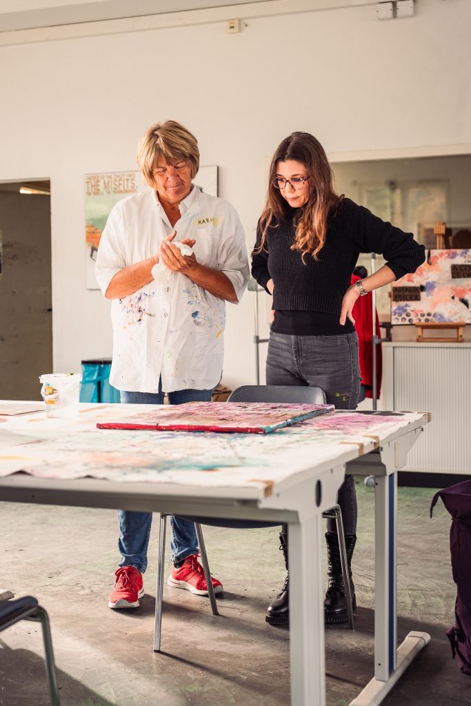Two women observe artwork on a table in an art studio one wearing a white shirt and the other in a black sweater