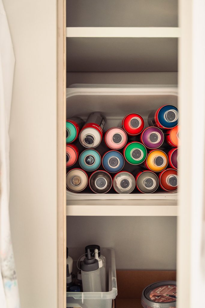 A shelf inside a cabinet displaying various spray paint cans in different colors and sizes organized with some additional art supplies like bottles and containers visible