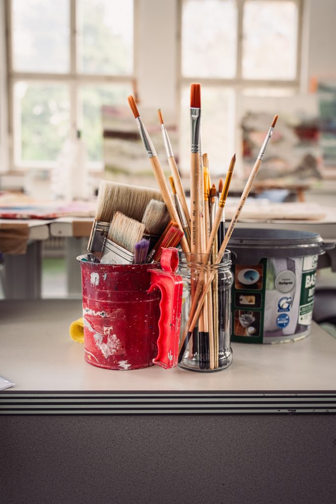 Painting with Julia | Atelier Siegen A collection of paintbrushes in a red container and a glass jar on a table in an art studio