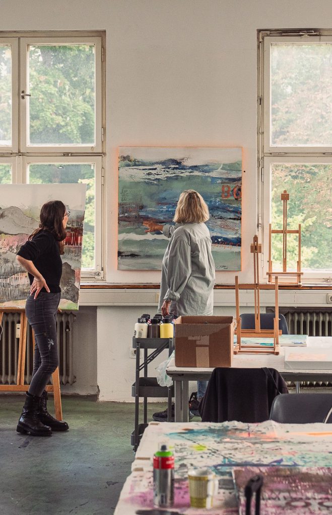 Two artists discussing artwork in a bright art studio with paintings on the walls and various art supplies on the tables