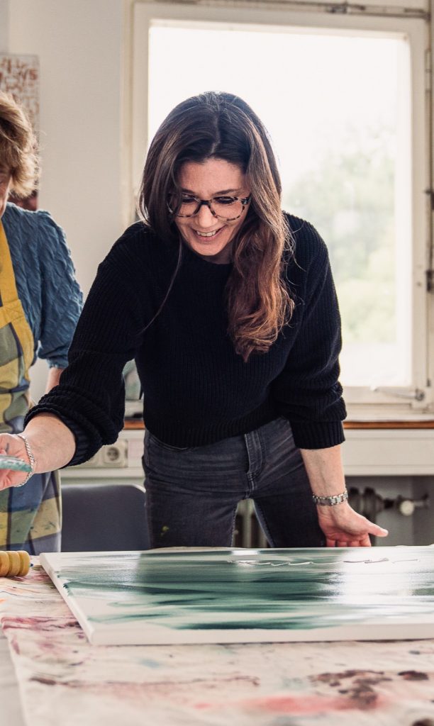 Julia Eckardt smiles while painting on a canvas showcasing a creative art studio environment with another person in the background