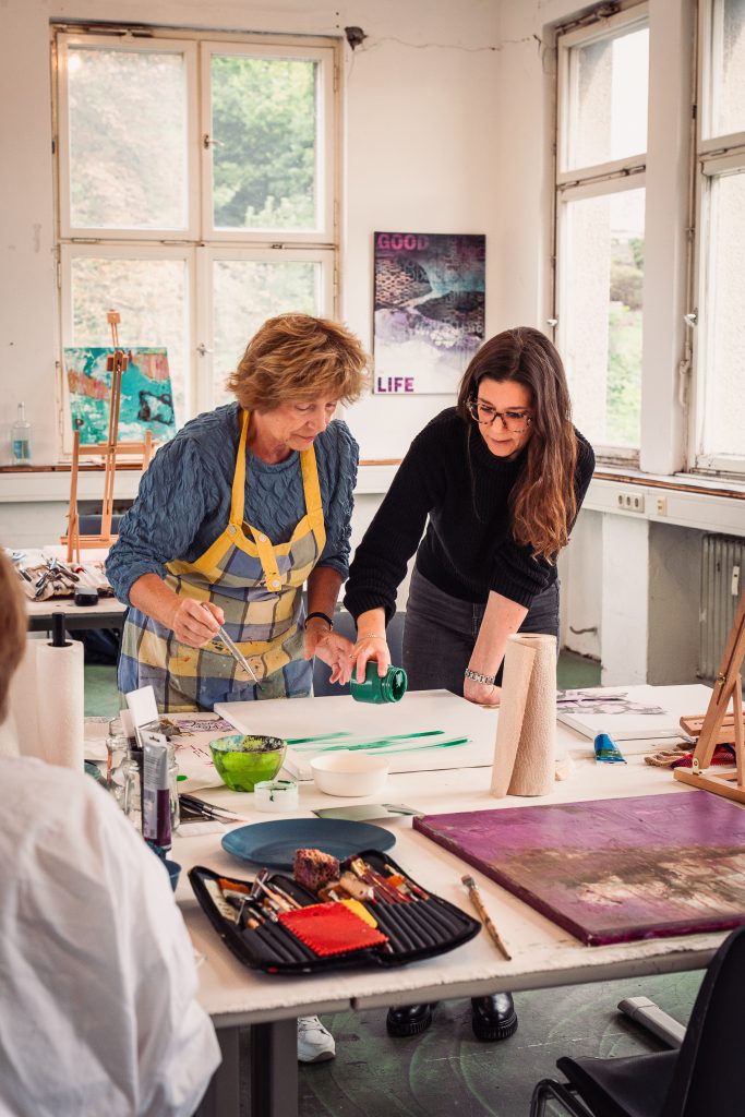 Painting with Julia | Atelier Siegen Julia Eckardt and a student engaged in an art class one teaching and the other learning The teacher is guiding the student Various art supplies are spread out on a table