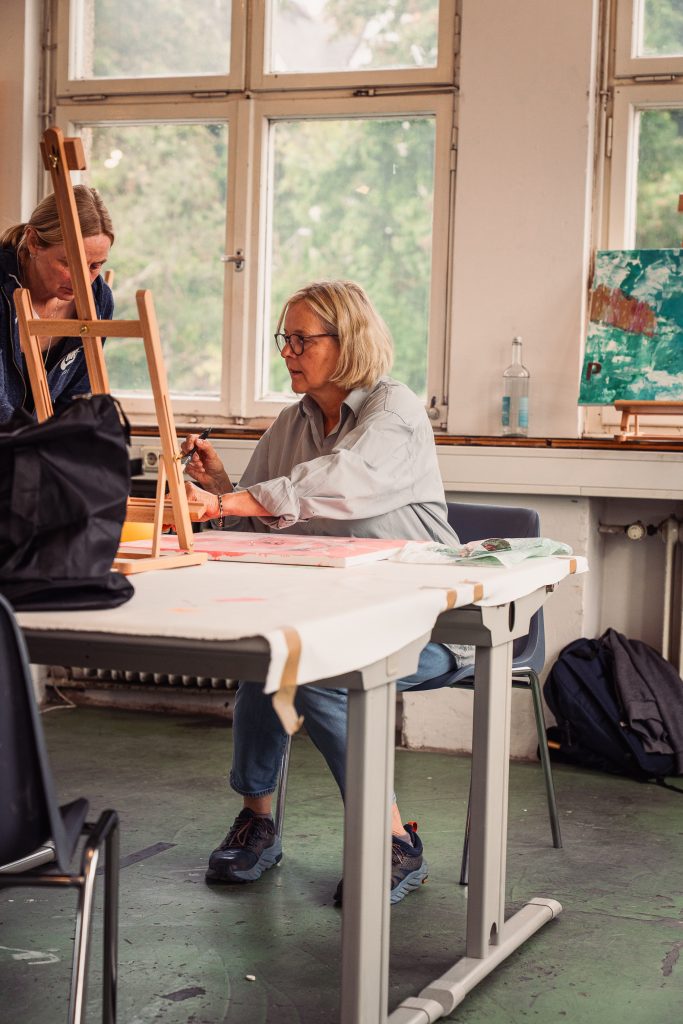 A woman is sitting at a table in an art studio focusing on her work Another person is leaning over an easel in the background while a colorful painting is visible on the wall