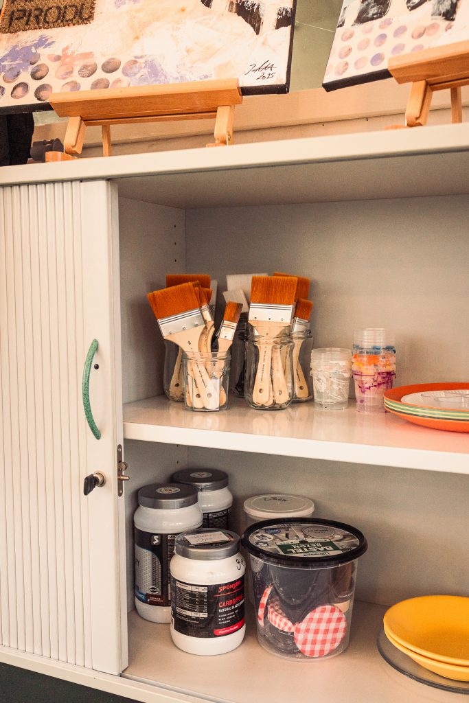 A storage cabinet with various art supplies including paintbrushes in jars and containers of paint The lower shelf features jars and packets along with colorful plates