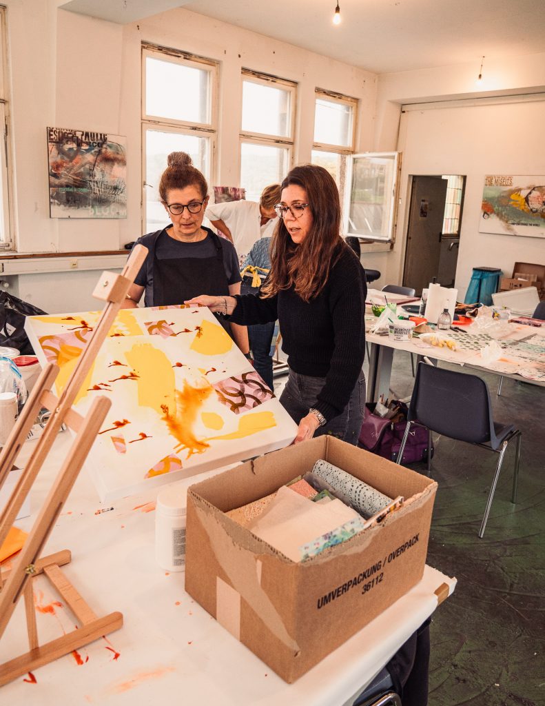 Two women discussing artwork in an art studio with one holding a brightly colored canvas An assortment of art materials and supplies are on a table