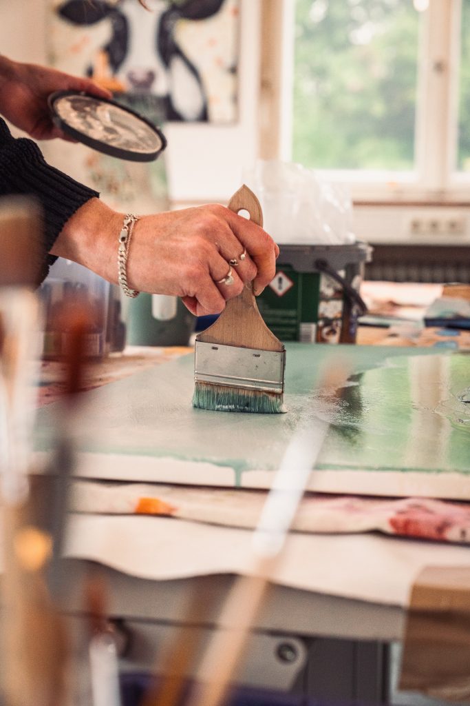 A person's hand holding a paintbrush, applying paint to a canvas in a bright and artistic workspace with various art supplies in the background.